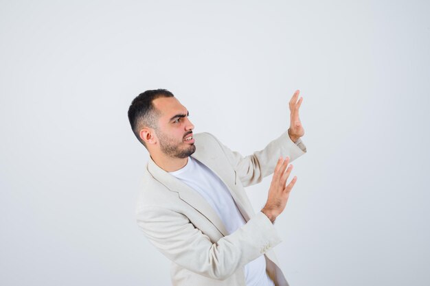 Young man stretching hands as stopping something in white t-shirt, jacket and looking scared. front view.