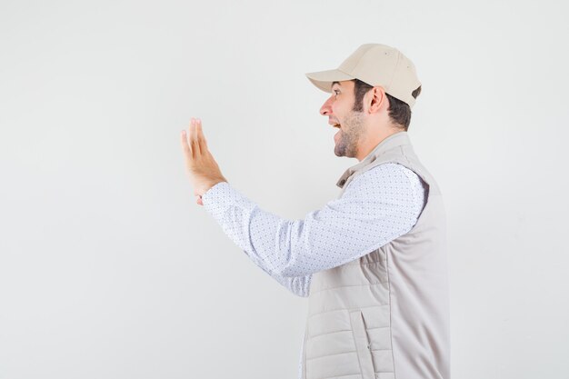 Young man stretching hands and laughing in beige jacket and cap and looking happy. front view.