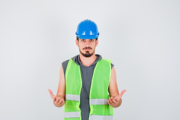 Young man stretching hands in questioning manner in construction uniform and looking happy