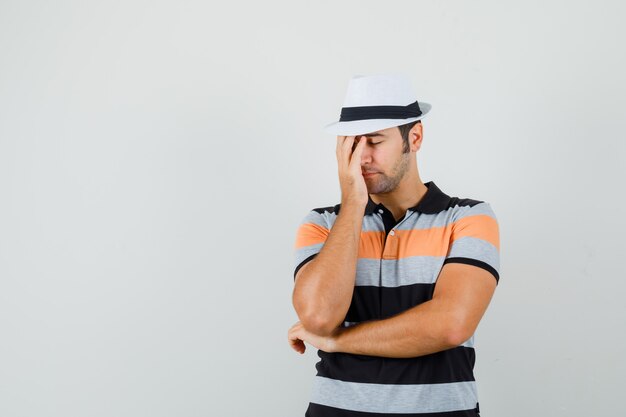 Young man in striped t-shirt,hat holding hand on his face and looking tired