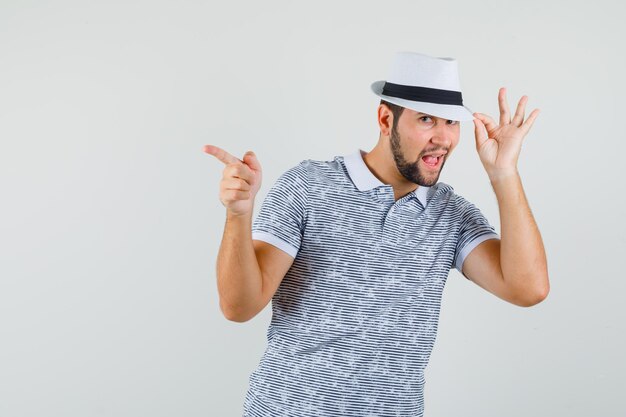 Young man in striped t-shirt,hat pointing away while touching at his hat and looking joyful , front view.