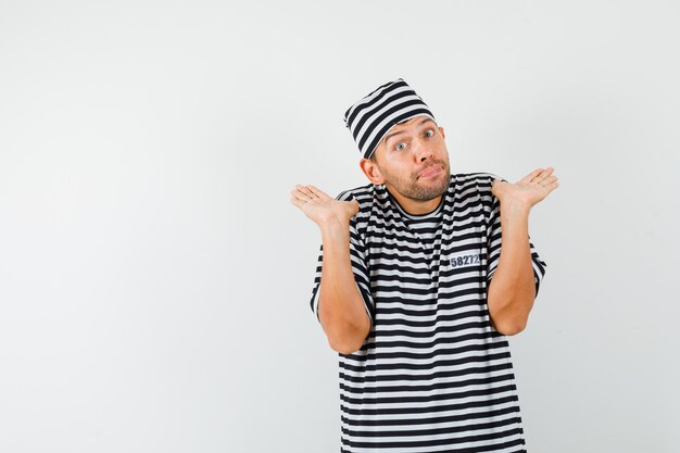 Young man in striped t-shirt hat showing helpless gesture by shrugging and looking confused  