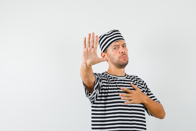 Young man in striped t-shirt hat showing stop gesture with hand on chest and looking annoyed  