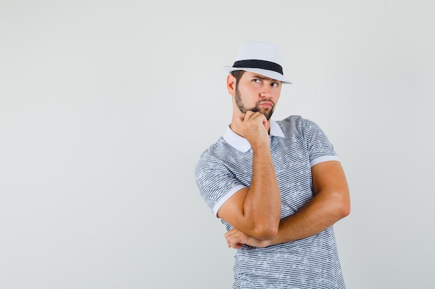 Молодой человек в полосатой футболке, шляпе, стоящей в позе мышления и озадаченной, вид спереди. место для текста