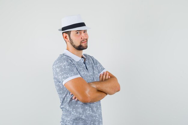 Young man in striped t-shirt,hat  while standing with crossed arms and looking confident , front view.