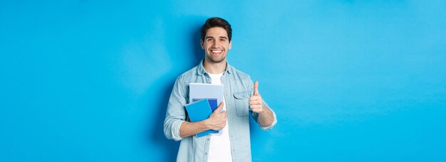 Young man student with notebooks showing thumb up in approval smiling satisfied blue studio backgrou