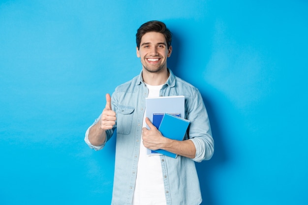 Young man student with notebooks, showing thumb up in approval, smiling satisfied, blue studio background