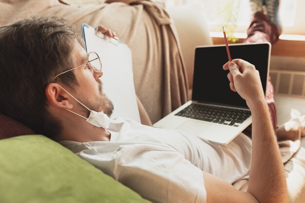 Young man studying at home during online courses for journalist, critics, writers. Getting profession while isolated, quarantine against coronavirus spreading. Using laptop, smartphone, headphones.