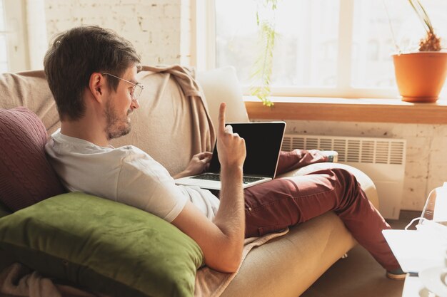 Young man studying at home during online courses for journalist, critics, writers. Getting profession while isolated, quarantine against coronavirus spreading. Using laptop, smartphone, headphones.