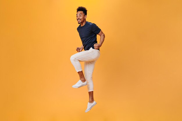 Young man in stylish outfit happily jumping on orange background Portrait of guy in white pants moving on isolated backdrop