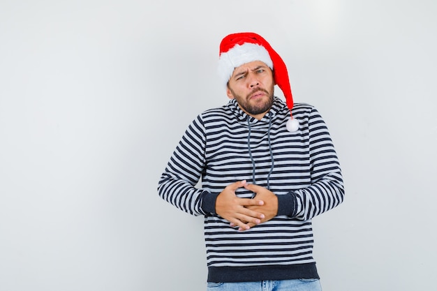 Young man suffering from abdominal pain in hoodie, Santa Claus hat and looking bothered , front view.