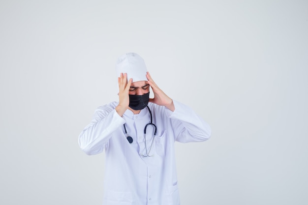 Young man suffering from headache in white uniform, mask and looking painful. 