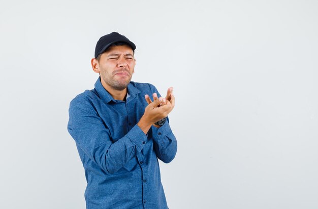 Young man suffering from painful hand in blue shirt, cap , front view.