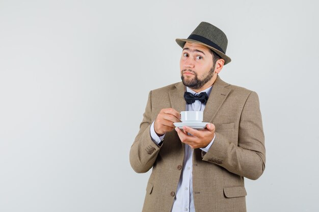 Young man in suit, hat drinking aromatic tea and looking positive , front view.