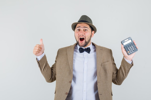 Молодой человек в костюме, шляпе, держа калькулятор с большим пальцем вверх и выглядя счастливым, вид спереди.