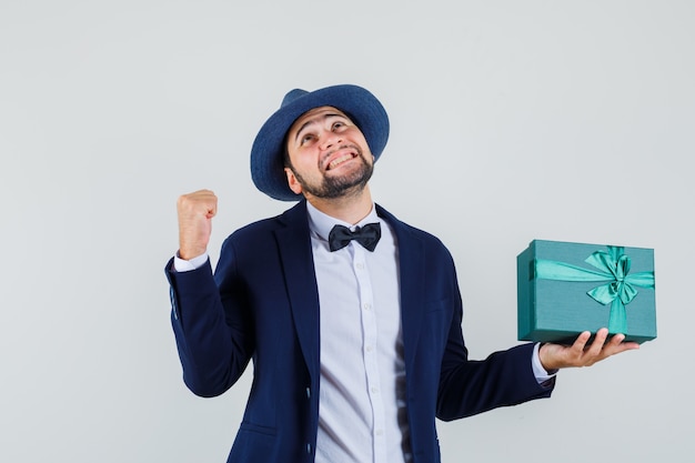 Молодой человек в костюме, шляпе, держащей настоящую коробку с жестом успеха и веселым взглядом, вид спереди.