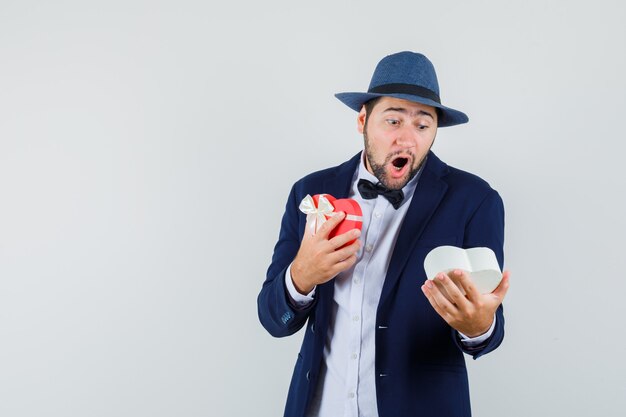 Young man in suit, hat looking into present box and looking surprised , front view.