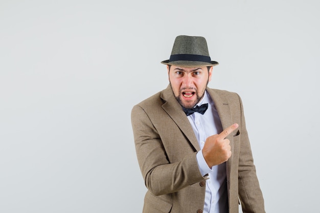 Young man in suit, hat shouting with finger up to the side and looking angry , front view.