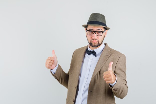 Молодой человек в костюме, шляпе показывает двойные пальцы вверх и выглядит уверенно, вид спереди.