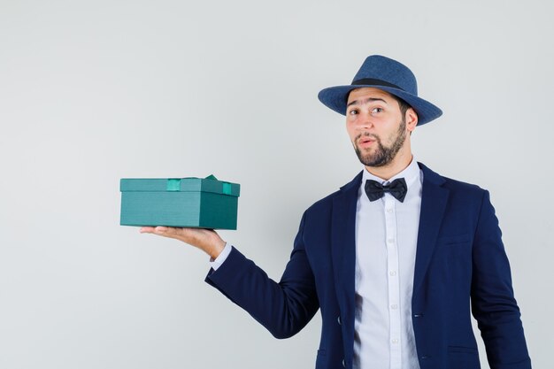 Young man in suit, hat showing present box and looking positive , front view.