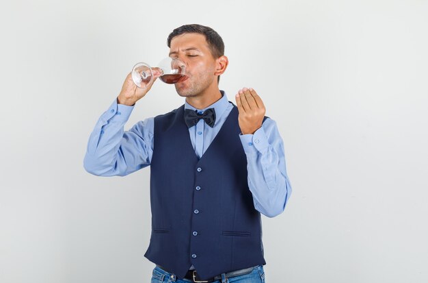 Young man in suit, jeans drinking alcohol and showing tasty gesture and looking pleased