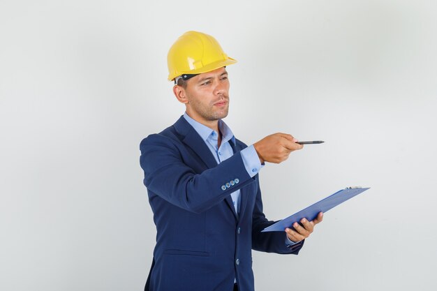 Young man in suit, safety helmet pointing away with pen and looking busy 