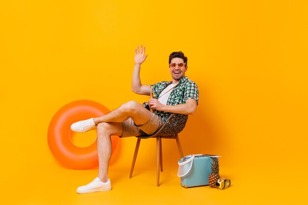 Молодой человек в солнечных очках наслаждается отпуском на фоне надувного круга и чемодана. Парень сидит на деревянном стуле, пьет пиво и машет рукой.