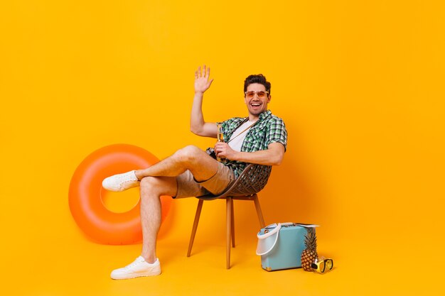 Young man in sunglasses enjoys vacation against backdrop of inflatable circle and suitcase. Guy is sitting on wooden chair, drinking beer and waving hand.