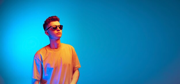 Молодой человек в солнечных очках и белой футболке позирует изолированно на синем фоне студии в неоновом свете