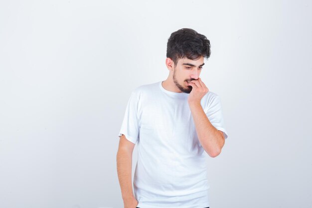 Young man in t-shirt biting his nails and looking concerned