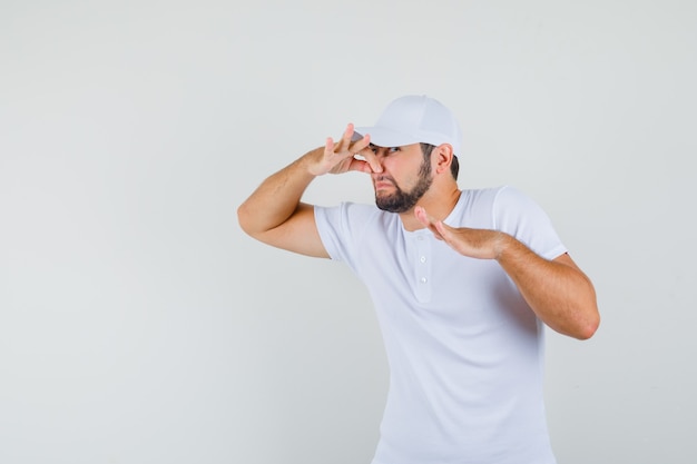 Young man in t-shirt,cap catching his nose and looking disgusting , front view.