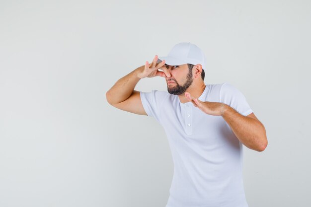 Young man in t-shirt,cap catching his nose and looking disgusting , front view.