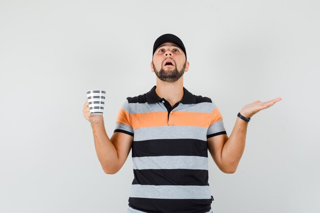 Young man in t-shirt, cap holding cup of drink, spreading palm aside and looking anxious , front view.