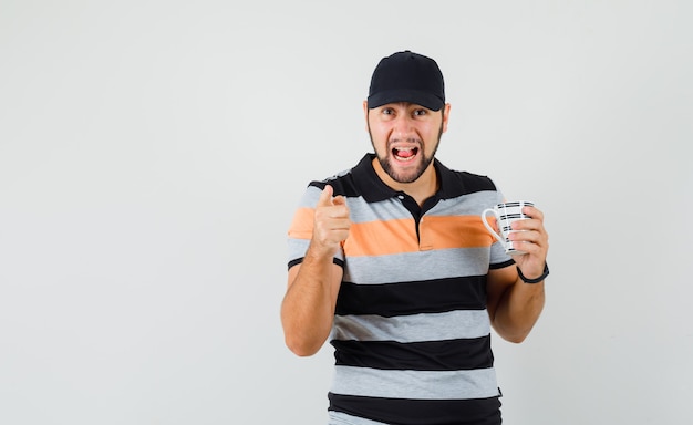 Young man in t-shirt, cap holding cup of drink, warning with finger and looking angry , front view.