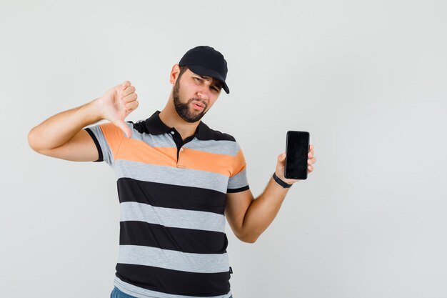 Young man in t-shirt, cap holding mobile phone, showing thumb down and looking disappointed , front view.