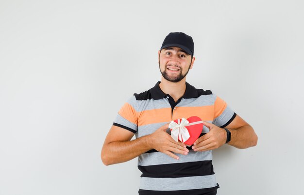 Young man in t-shirt, cap holding present box and looking merry , front view.