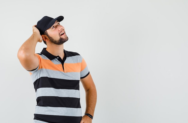 Foto gratuita giovane uomo in t-shirt, berretto alzando lo sguardo con la mano sulla testa e guardando sognante, vista frontale.