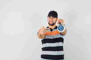 Free photo young man in t-shirt, cap pointing at alarm clock and looking confident