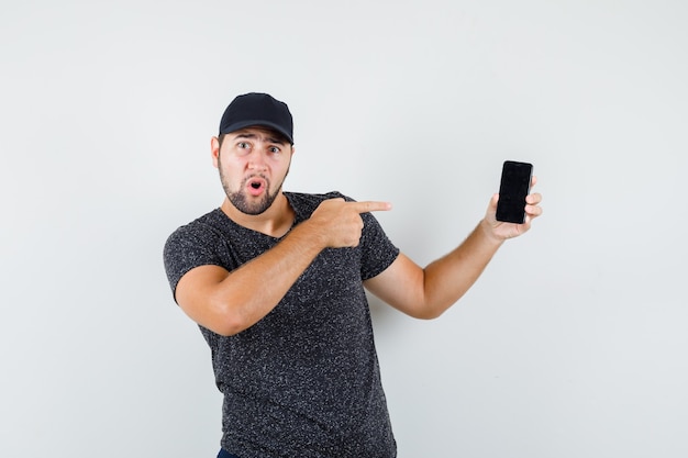 Young man in t-shirt and cap pointing at mobile phone and looking confident