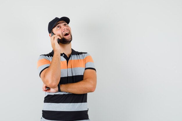 Young man in t-shirt, cap talking on mobile phone and looking happy , front view.
