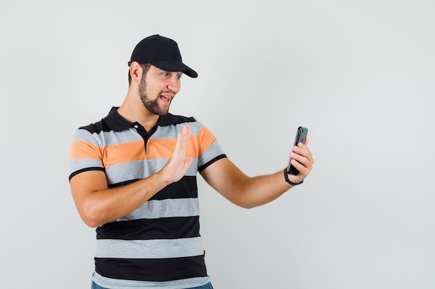 Young man in t-shirt, cap waving hand on video chat and looking cheerful , front view.