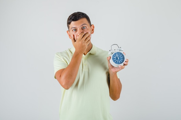 Young man in t-shirt covering mouth and holding alarm clock and looking surprised