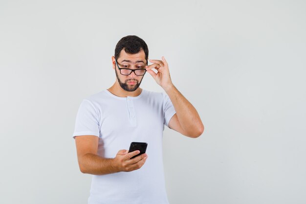 Young man in t-shirt,glasses looking at phone and looking careful , front view.