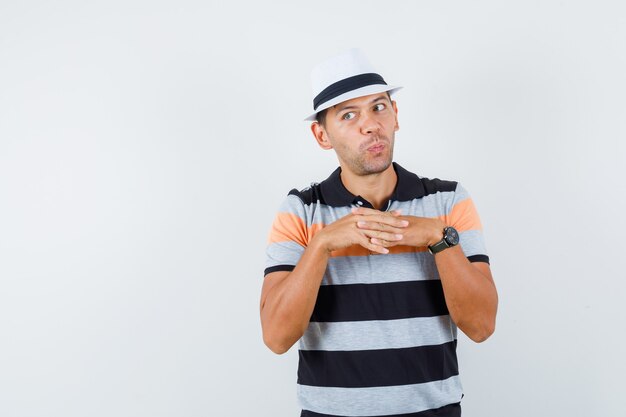 Young man in t-shirt and hat bridging fingers, looking away and looking curious