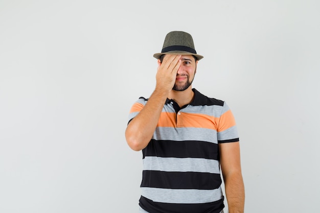 Young man in t-shirt, hat holding hand on one eye and looking ashamed , front view.