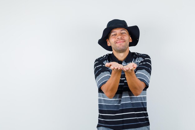 Young man in t-shirt, hat holding palms together and looking cheerful , front view.