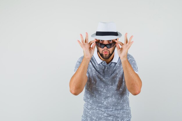 Young man in t-shirt,hat looking over his sunglasses and looking surprised , front view.