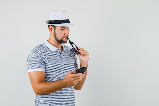 Tシャツを着た青年、眼鏡をかけながらスマートフォンを見て物思いにふける帽子、正面図。