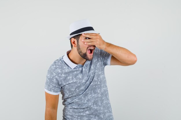 Young man in t-shirt, hat looking through fingers and looking curious , front view.