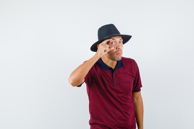 Young man in t-shirt, hat opening eye with fingers, front view.